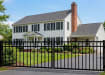 A decorative black Aluminum Driveway Gate pair with a 3-rail staggered spear design installed across an asphalt driveway leading to a large white traditional-style home.